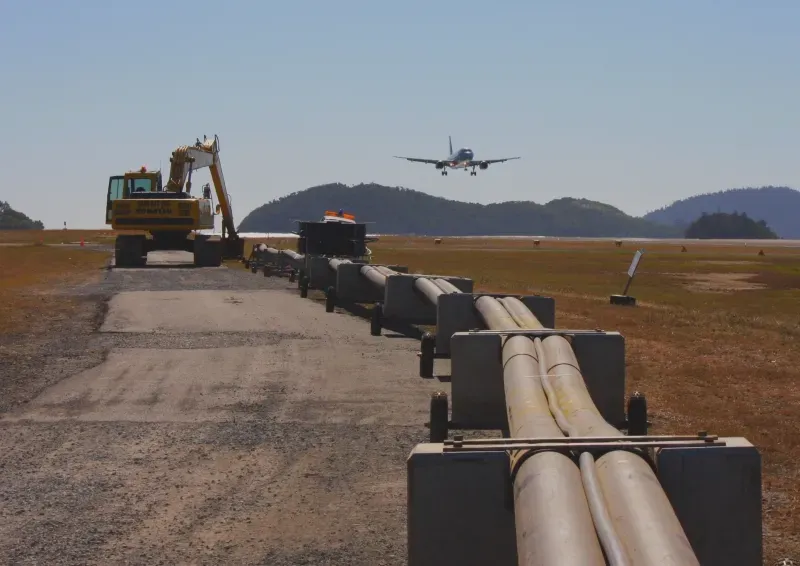 Submarine fibre pipeline bundle staged on concrete cradles along the active Hamilton Island airstrip, with an aircraft on final approach landing in the background. Excavator at the rear of the bundle.