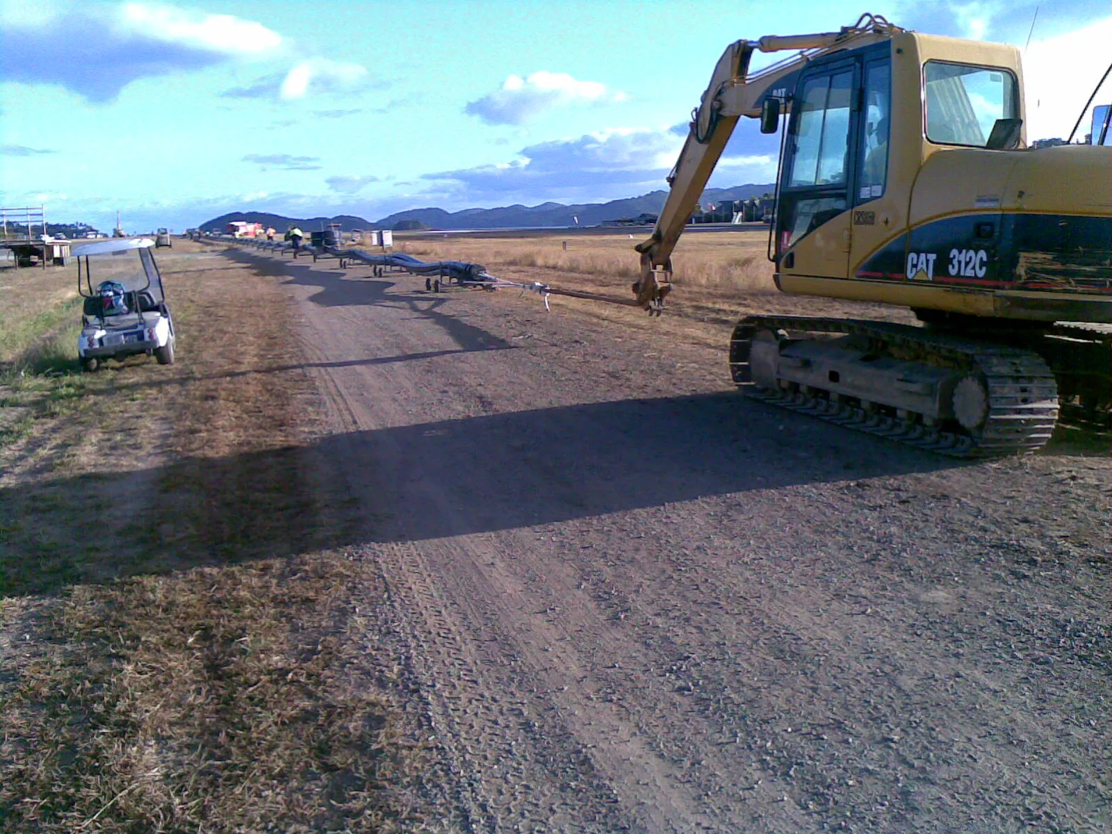 Caterpillar 312C excavator alongside the staged pipeline on the Hamilton Island airstrip. Pipeline on rolling cradles receding into the distance toward the worksite, with a golf cart at the apron edge.