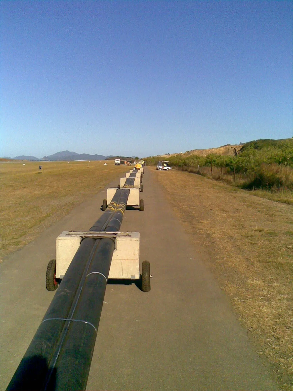 Submarine pipeline bundle on rolling concrete cradles, lined up along the sealed apron beside the Hamilton Island airstrip. Crew and equipment in the distance.