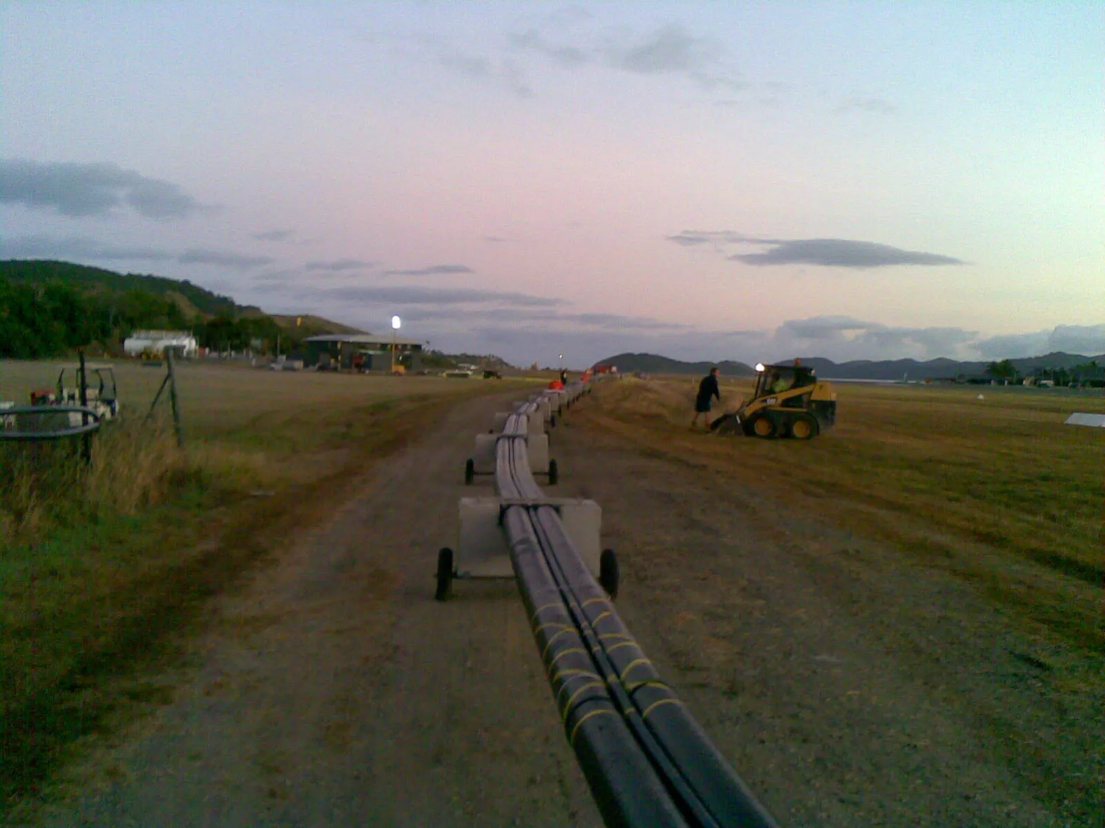 Submarine pipeline staged along the Hamilton Island airstrip apron at dusk. Skid steer working alongside, operations buildings in the distance under a Whitsunday sunset sky.