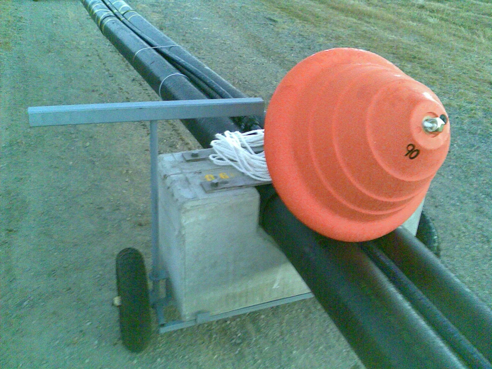 Close-up of a rolling cradle supporting the pipeline bundle. Orange pipe end cap, white deployment rope coiled on the concrete weight, twin HDPE pipes carrying the submarine fibre conduit.