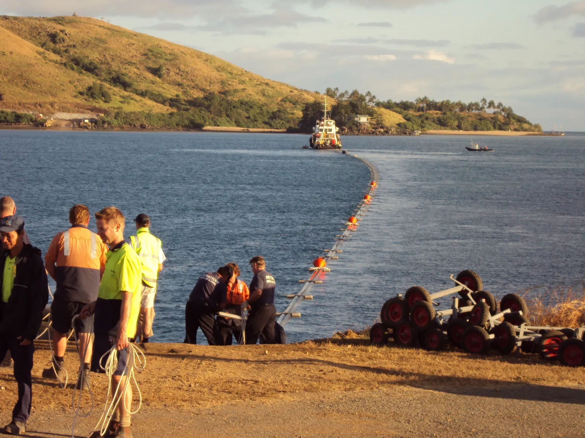 Crew gathered at the Dent Passage shoreline as the pipeline is fed from the Hamilton Island airstrip into the water. Skid steer on the right, pipeline on rolling cradles in the foreground, Whitsunday islands across the bay.