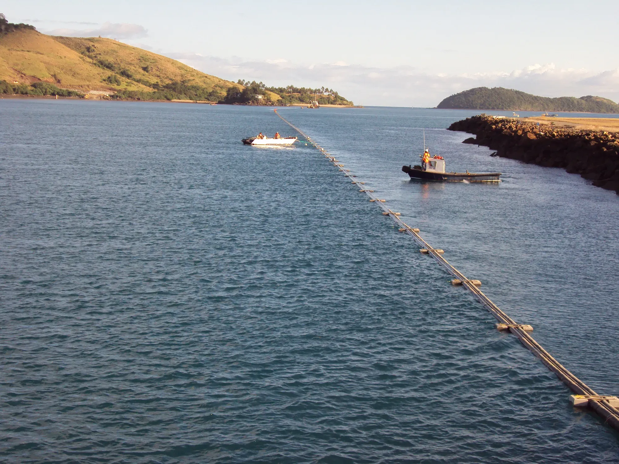Submarine fibre pipeline bundle being towed across Dent Passage from Hamilton Island, marked with surface floats. Accompanying vessels mid-tow and tugboat at the landfall. Whitsunday hills in the background.