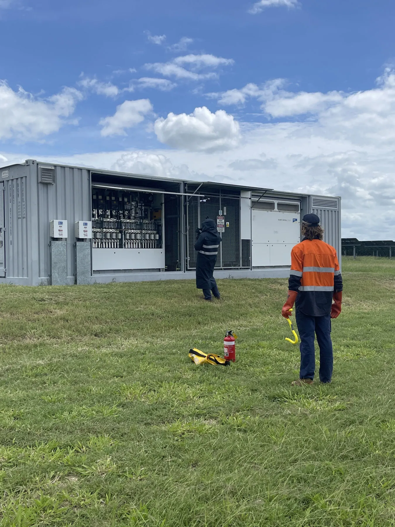 Two technicians in hi-vis conducting shutdown and safety procedures at an open inverter station, Hayman Solar Farm, Queensland — fire extinguisher on standby.